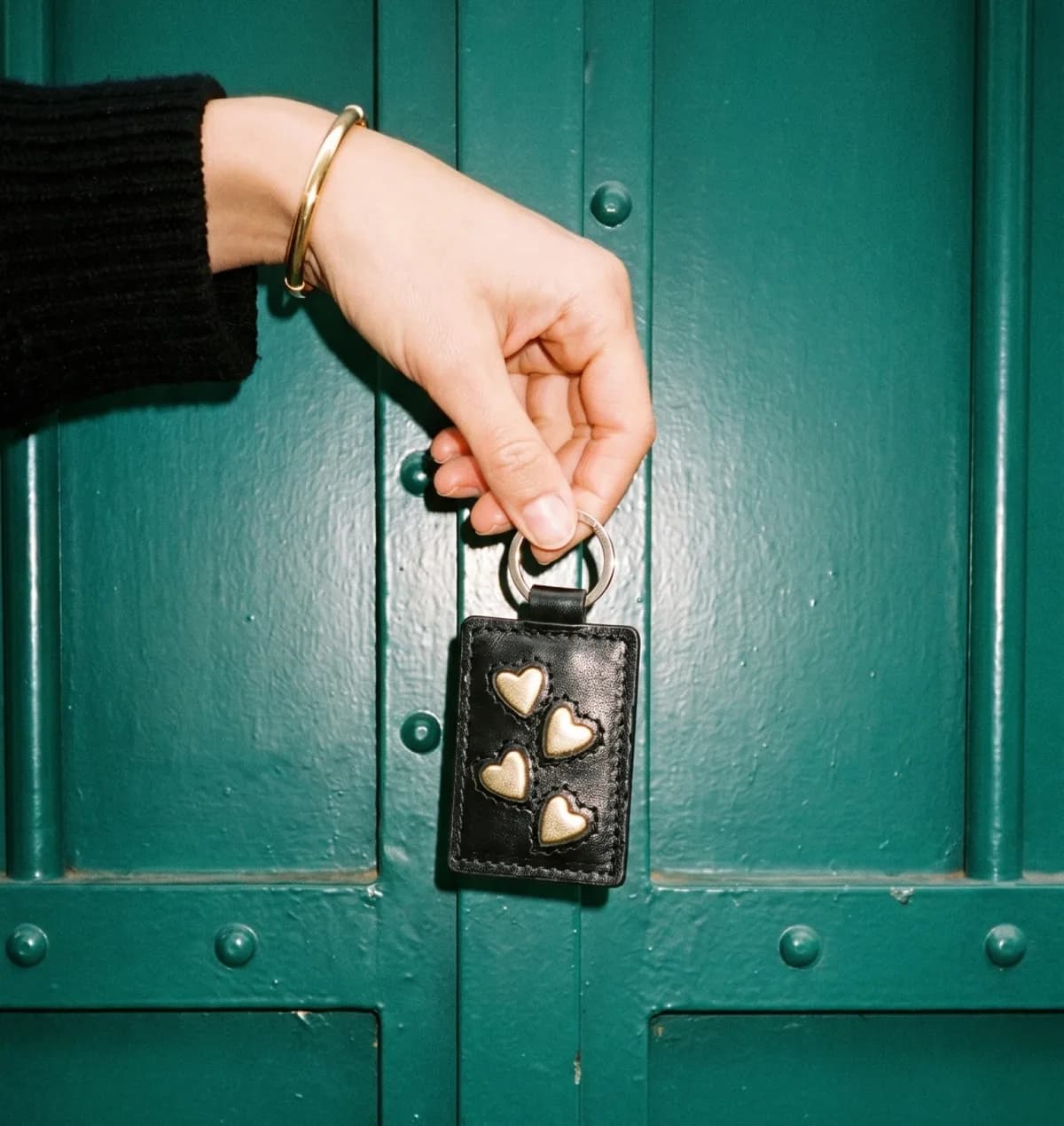 Porte-clé en cuir noir aux cœurs dorés tenu devant une porte parisienne verte — Suki Paris