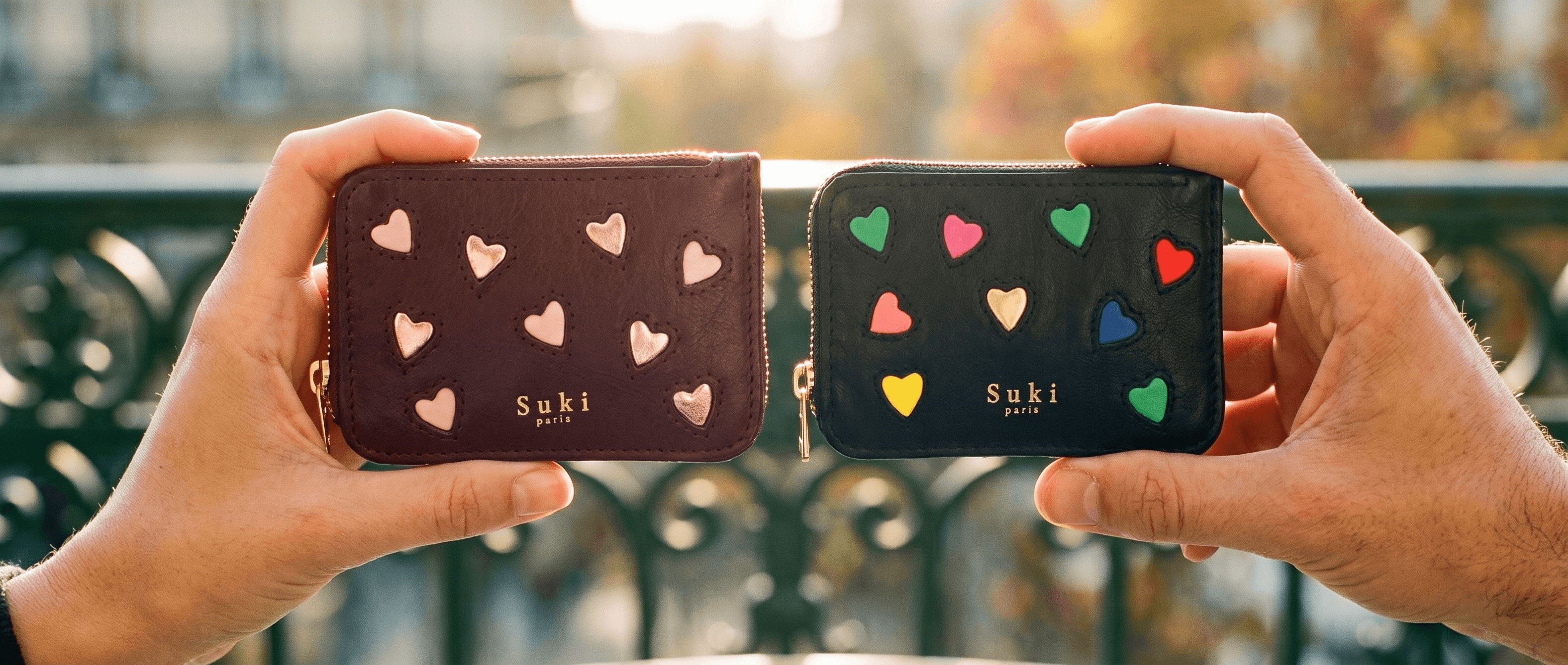 Deux porte-monnaies en cuir à cœurs Suki Paris, bordeaux et noir, tendus l'un vers l'autre sur une terrasse parisienne